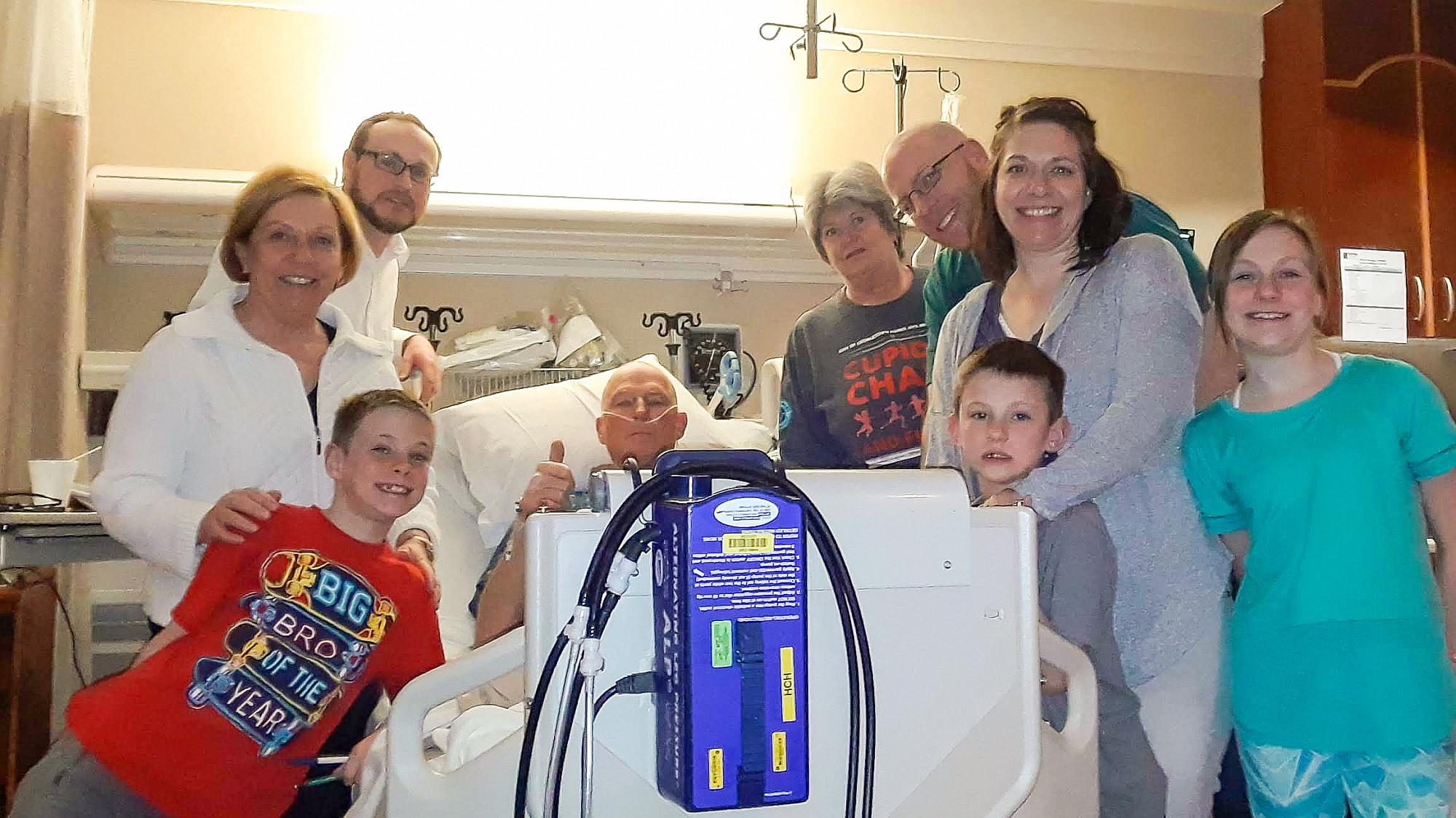 The Simonson family rallies around Jerry after his surgery. Jerry lies in a hospital bed, giving a thumbs up, while his family stands around the bed and smiles for the camera.