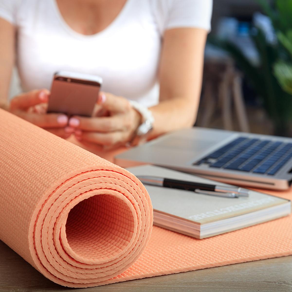 A peach yoga mat rests on a table next to an open laptop. A woman's hands hold a cell phone in the background.