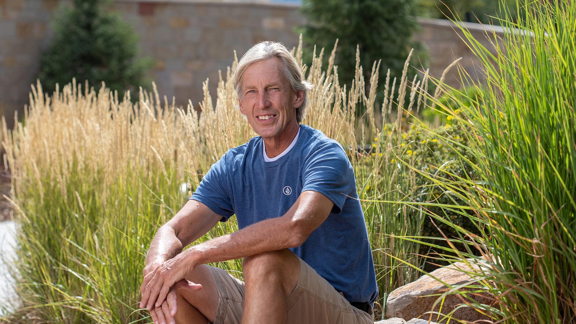 Randy Oldham, an older male patient with silver hair sits outside on a rock with tall grasses behind him.
