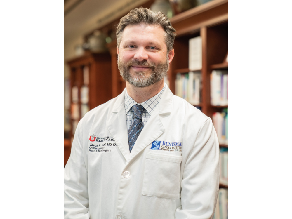 White man names Jason Hunt wearing a white lab coat over a white button up and blue tie standing in front of bookshelves