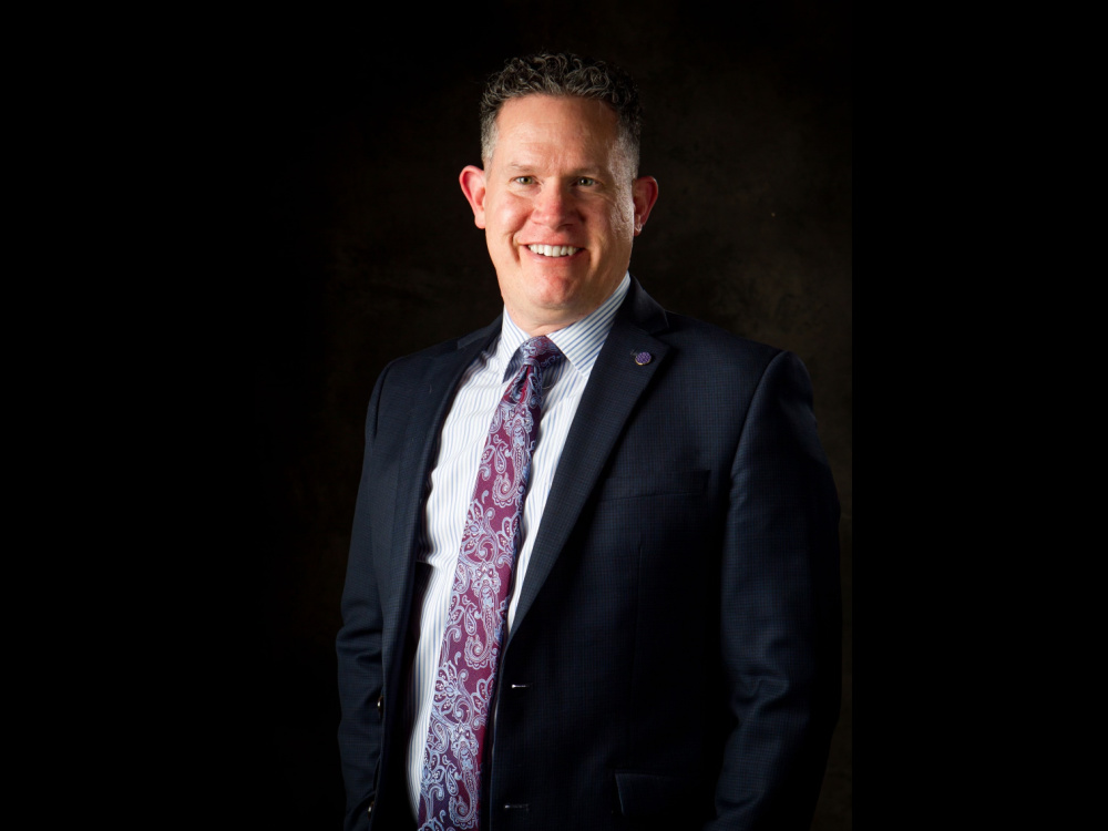 White man named Loren Savage wearing dark suit jacket over a light grey shirt and purple tie