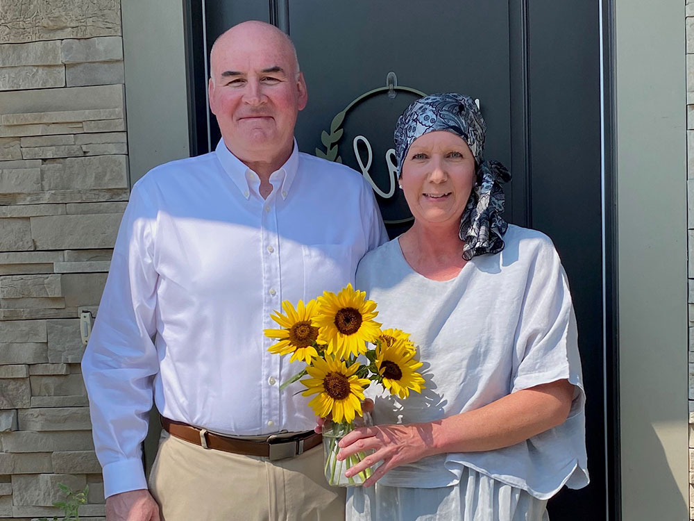 Pam wearing a head scarf and holding flowers, standing next to her husband