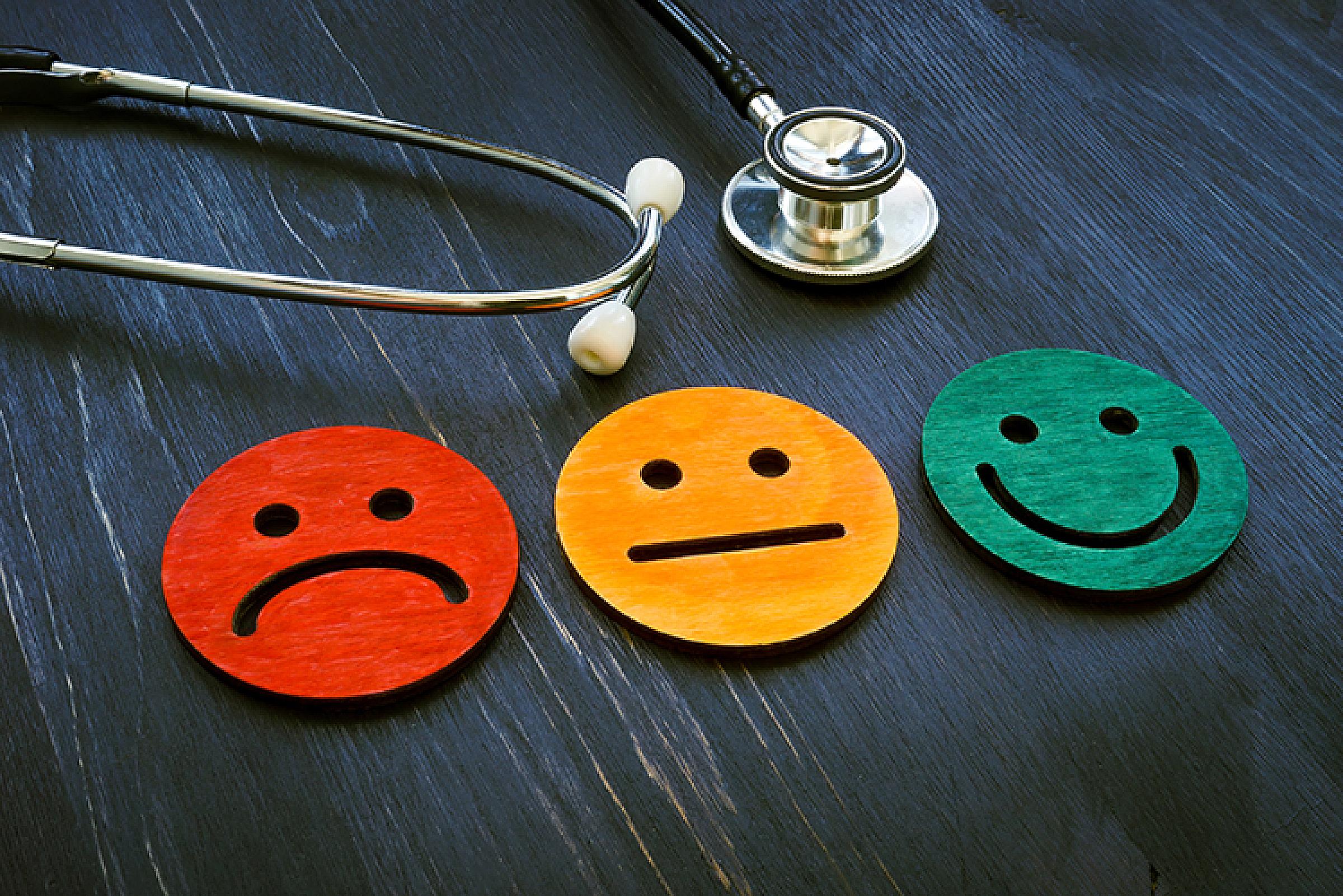 Red disk with frowning face, yellow disk with neutral face, and green disk with smiling face on a table next to a stethoscope