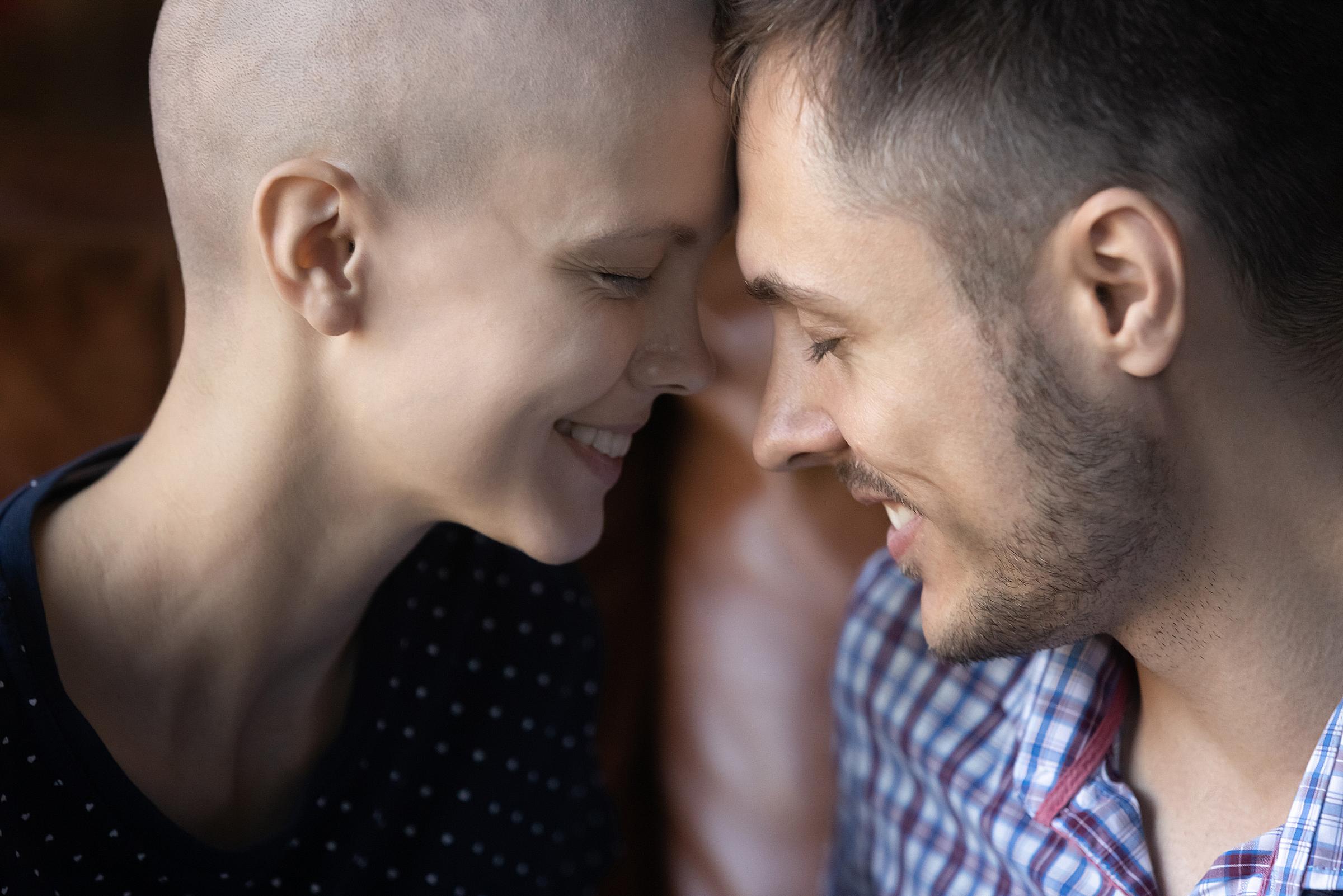 A couple lean foreheads together while smiling, one is bald from chemo treatments
