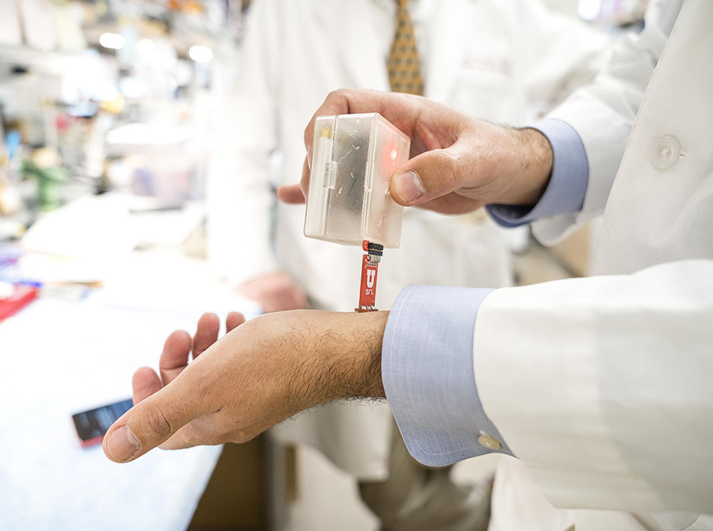 Doctor holding a URKSIN device over his wrist; the device is a small rectangular box with a red tab extended from on one end to press against the skin
