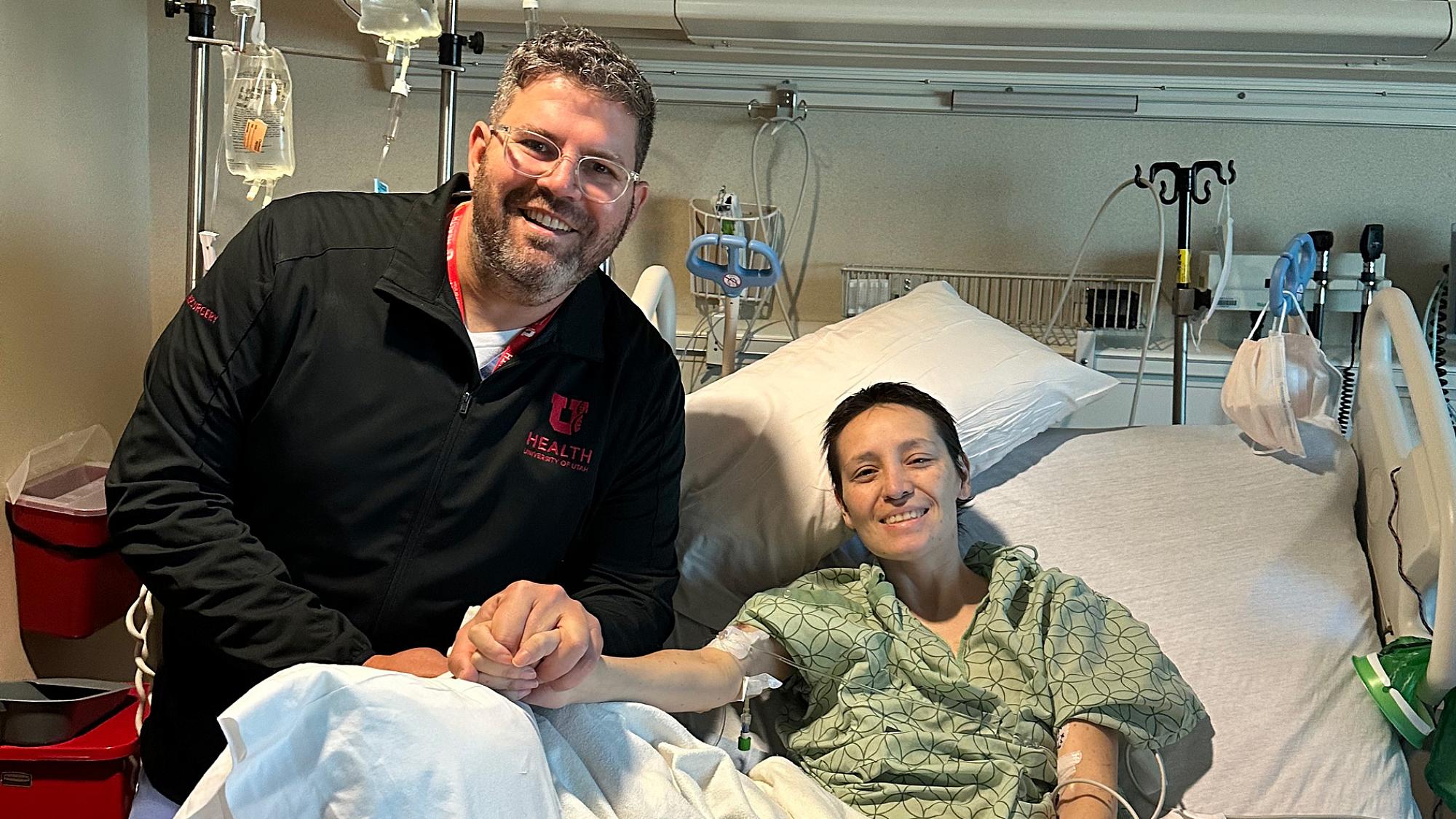 Ben Shofty, MD, PhD, and Natalia Garcia holding hands while she lies in a hospital bed and smiles at the camera