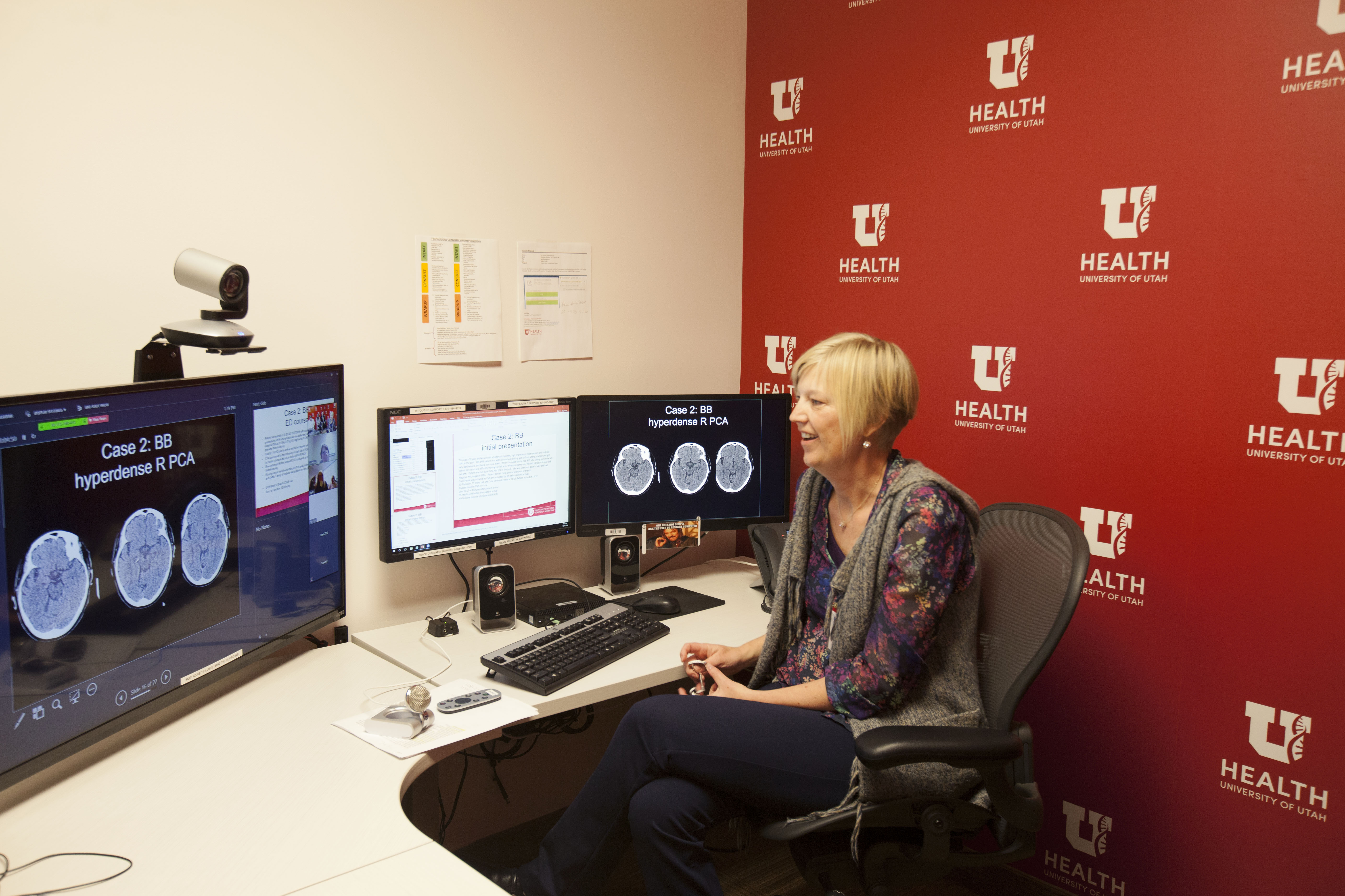 U of U Health female stroke provider sits in office chair talking to patient via video with brain scans on computer screens in the background