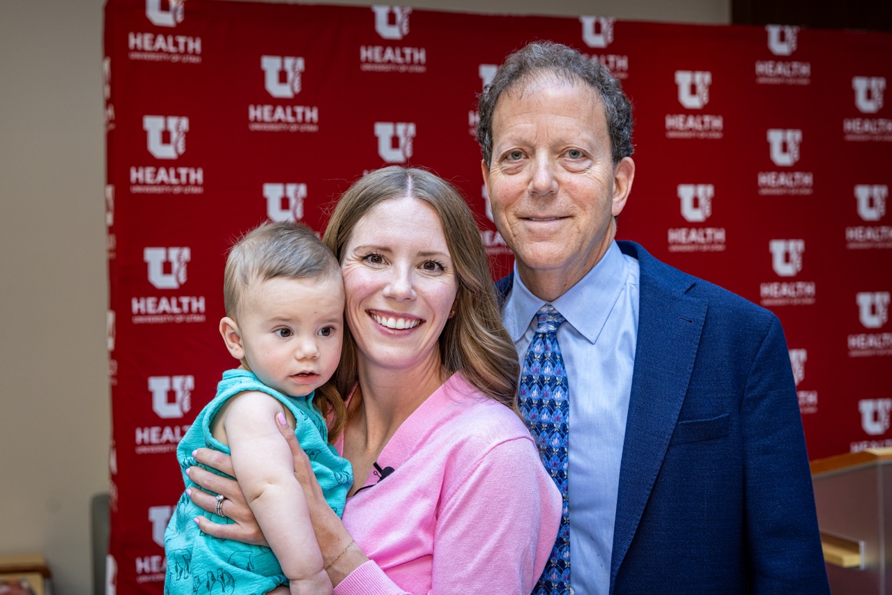 A smiling woman holds a baby, standing next to a man in a white coat, at the ribbon cutting ceremony for the University of Utah Health Pregnancy After Loss Program.