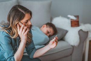 Parent looking at thermometer while on phone with child in background.