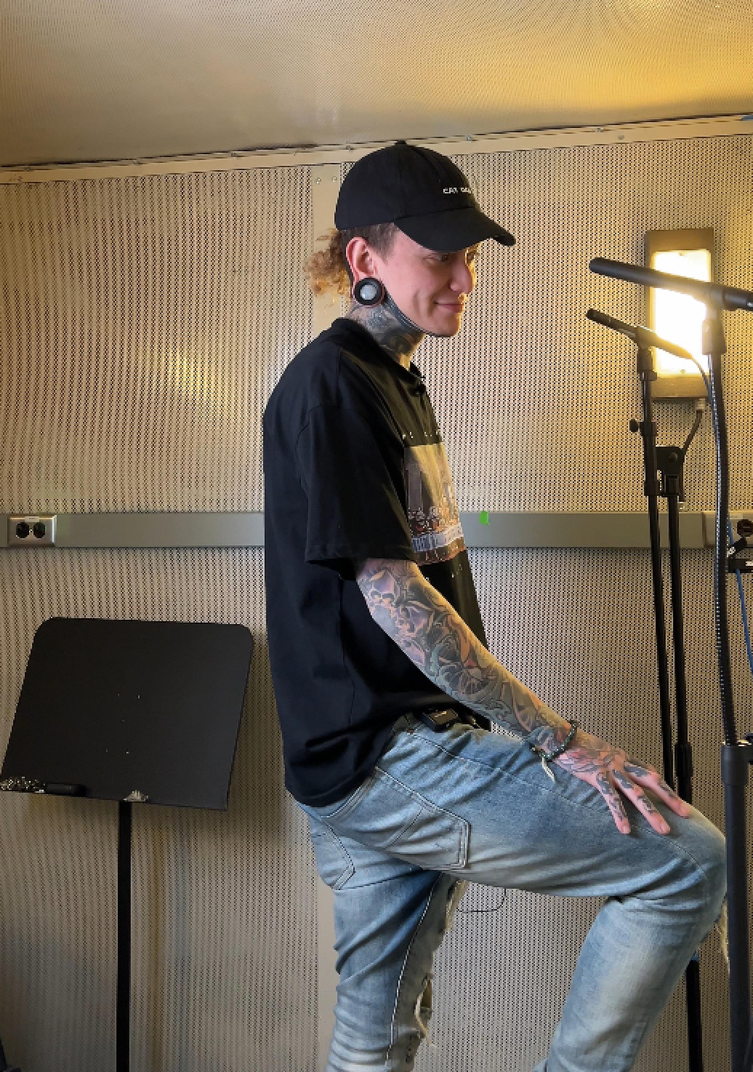 A person with full sleeve tattoos cups his ears as he sings in a recording booth