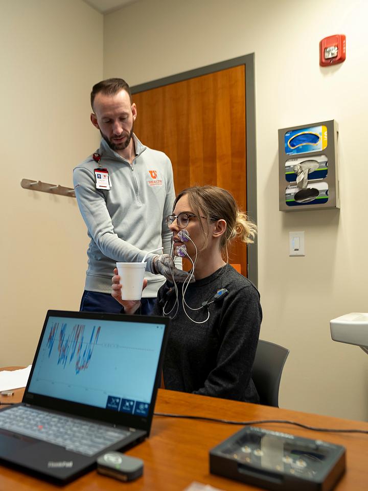 A U of U Health provider helps a woman drink from a straw. Her facial muscles are connected to nodes and seen on a screen in the background.