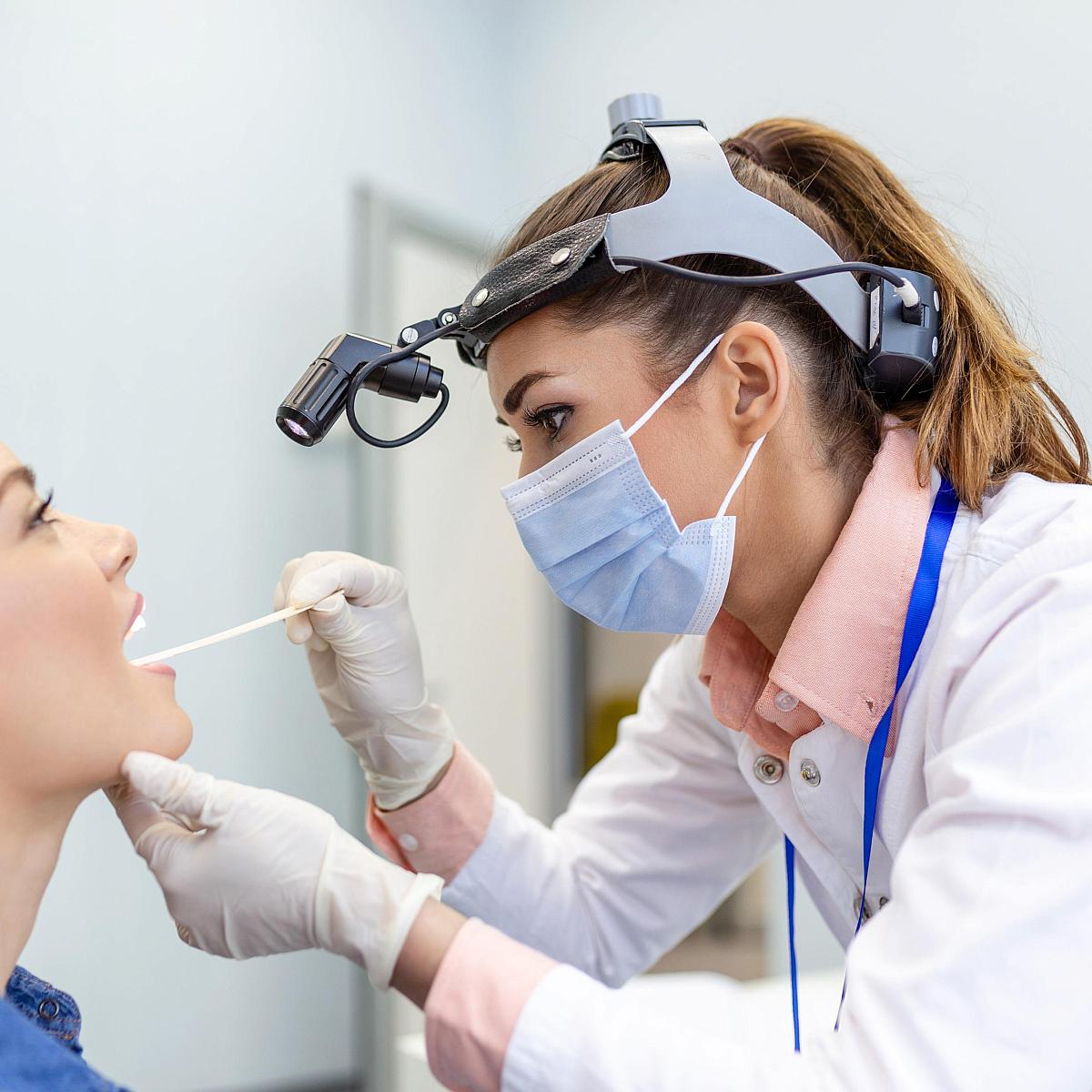 Picture of white female doctor wearing a face mask while she swabs a young female patient's throat.