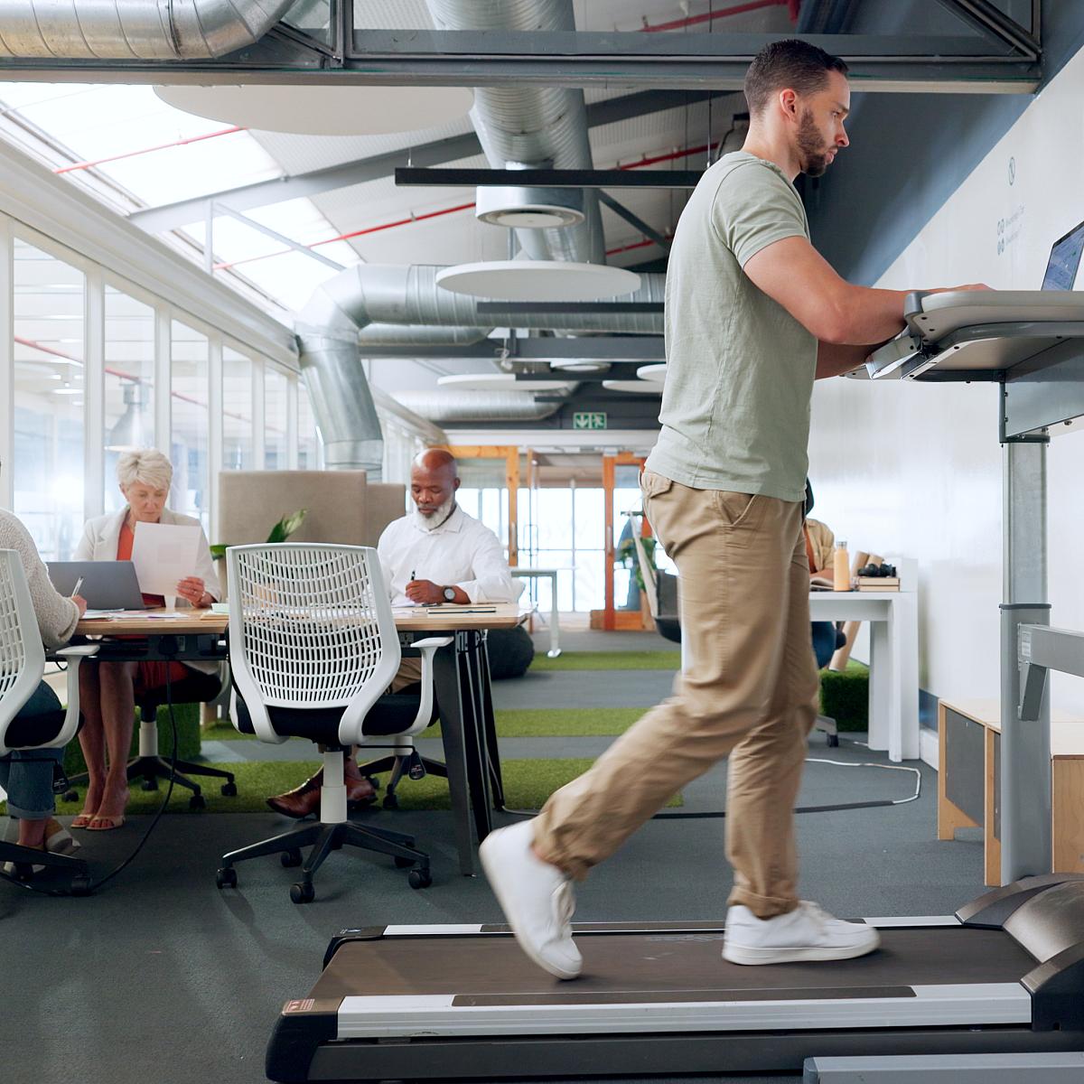 Man works at a treadmill desk while three people sit at a table behind him.