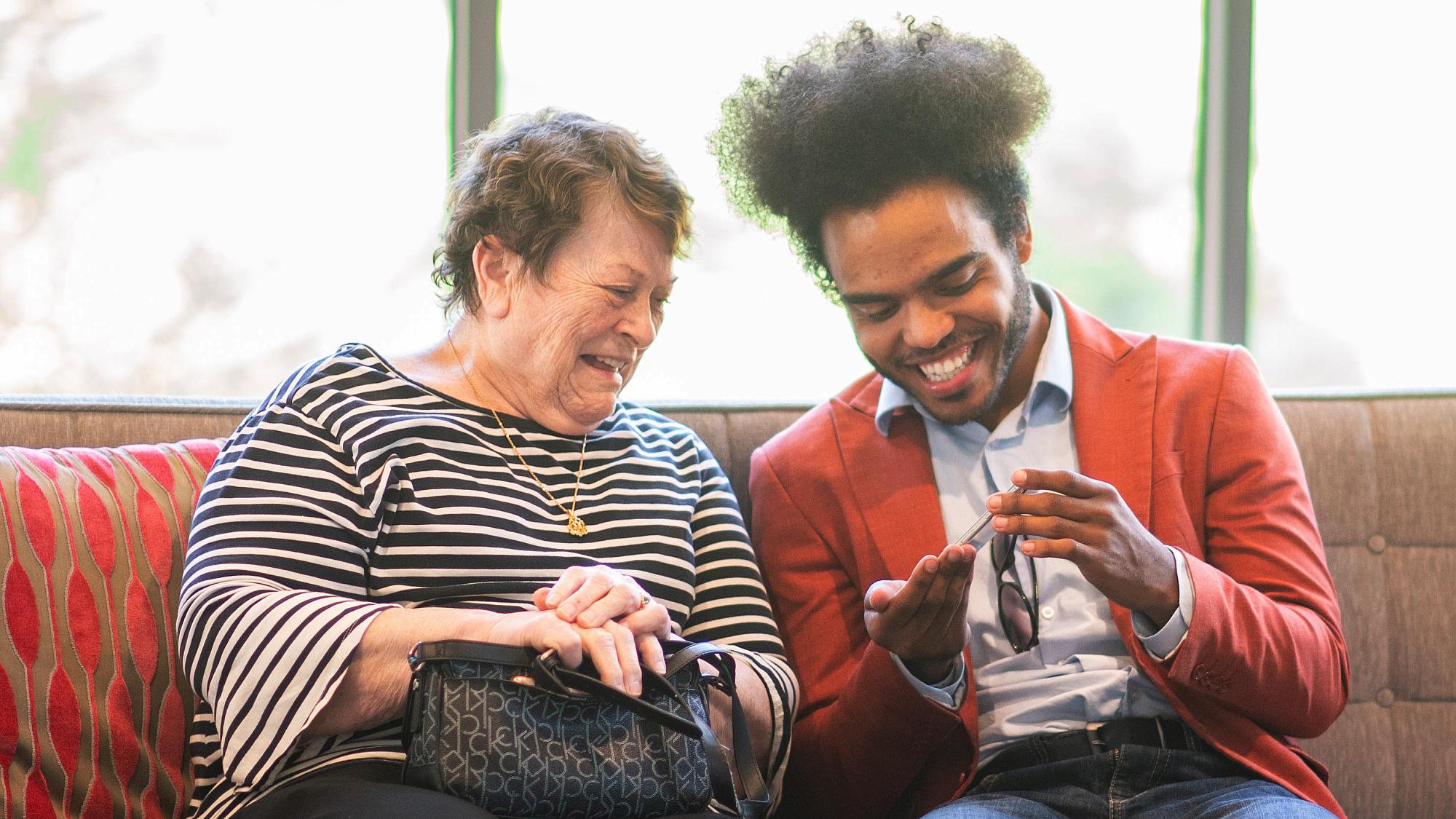 Older adult woman sitting on couch with young man both smiling looking at something on his phone