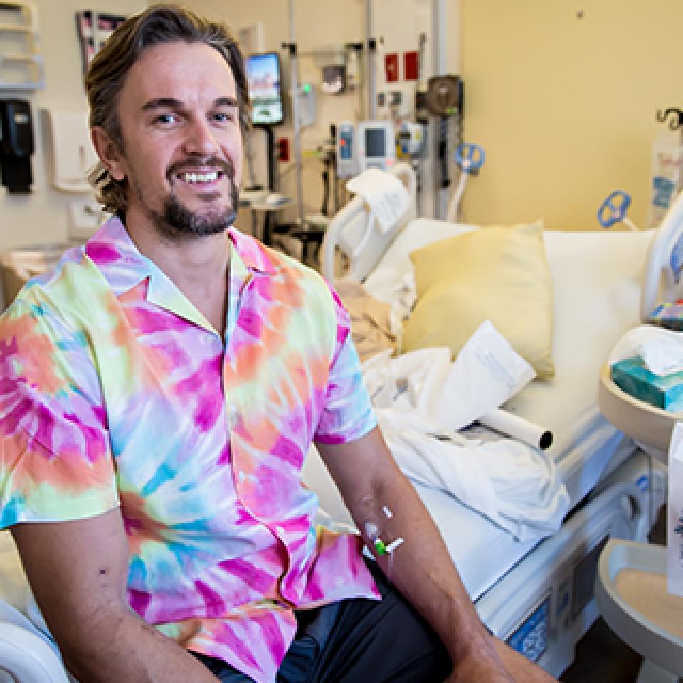 Stroke patient, Mark Kroeze, a man with light skin sits on a hospital bed and wears a tie dye shirt.